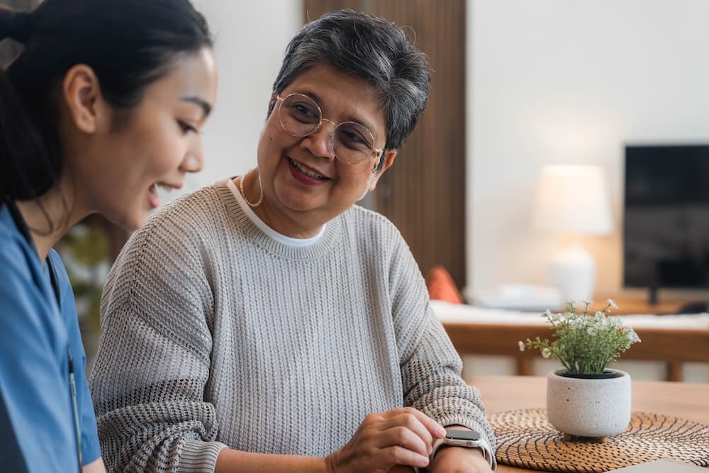 A young female caregiver in scrubs smiles while looking at an older woman with glasses, who checks her smartwatch while seated at a table.