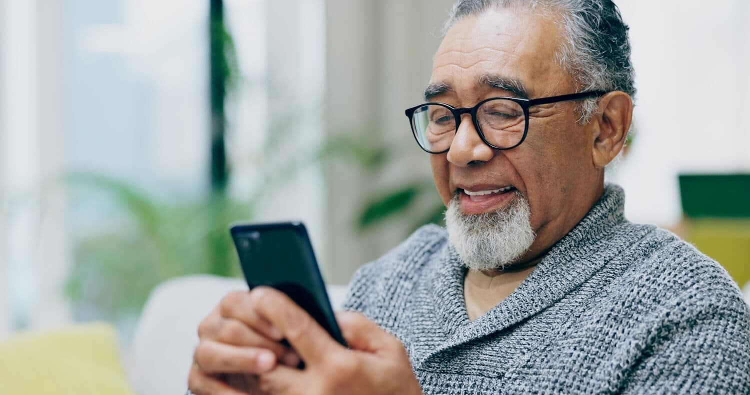 An older man smiles while looking at a smartphone, wearing a gray sweater.